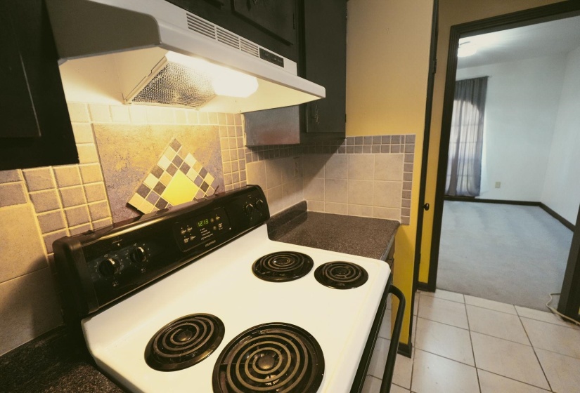 Kitchen with ventilation hood, white electric stove, light colored carpet, dark countertops, and decorative backsplash