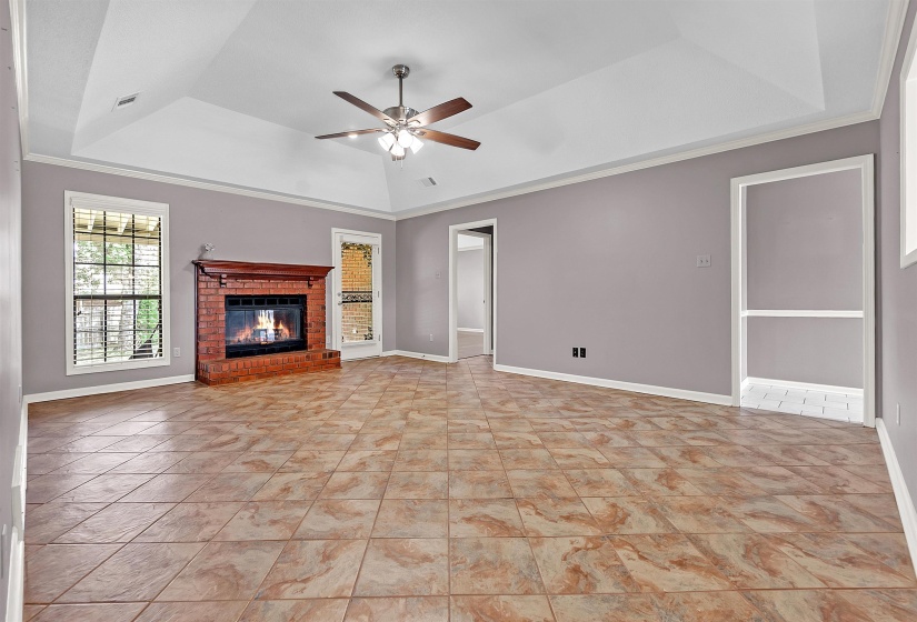 Unfurnished living room featuring a tray ceiling, ceiling fan, a brick fireplace, crown molding, and light tile patterned floors
