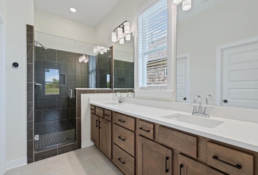 Bathroom featuring a glass-enclosed shower with dark tile surround, a dual vanity with white countertops, and wood-finish cabinetry with dark hardware