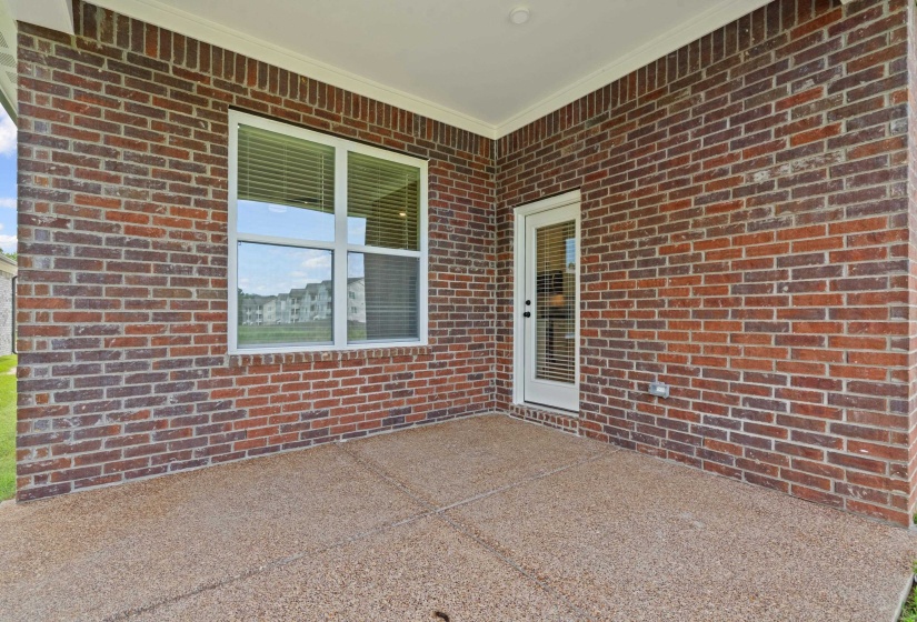 Covered patio featuring brick exterior, exposed aggregate concrete flooring, recessed lighting, a multi-pane window with blinds, and a glass-paneled exterior door