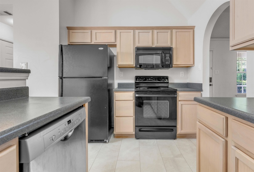 Kitchen featuring black appliances, light wood finish cabinets, dark countertops, arched walkways, and light tile patterned flooring