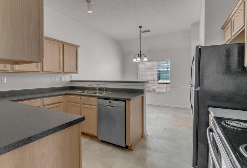 Kitchen with dark countertops, light wood finish cabinetry, stainless steel appliances, hanging lights, and a peninsula
