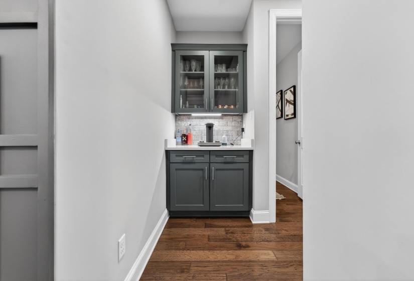Indoor dry bar with gray cabinetry, glass fronted cabinets, dark wood-style flooring, decorative backsplash, and light stone counters