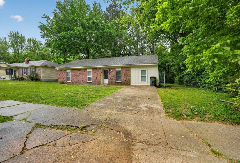 Ranch-style house with a front lawn, brick siding, concrete driveway, a shingled roof, and view of scattered trees