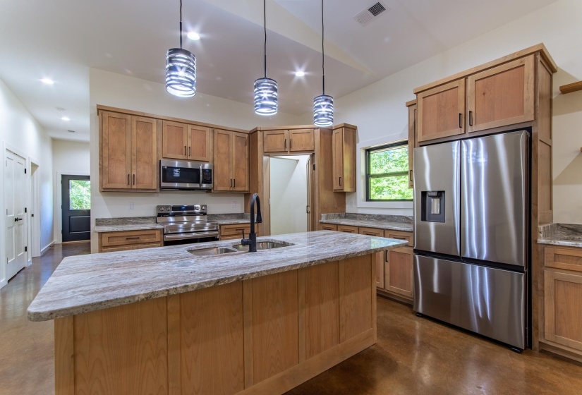 Kitchen featuring light stone countertops, concrete flooring, stainless steel appliances, an island with sink, and decorative light fixtures