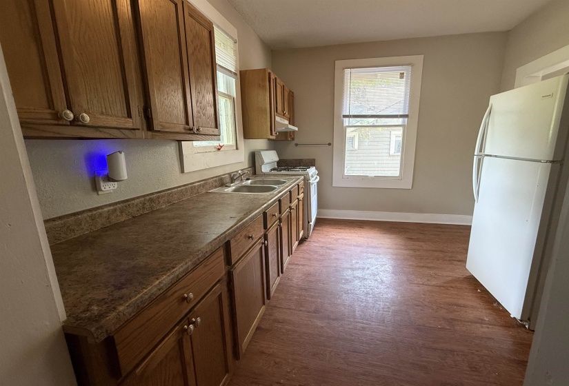 Kitchen with white appliances, dark wood-type flooring, brown cabinetry, dark countertops, and under cabinet range hood