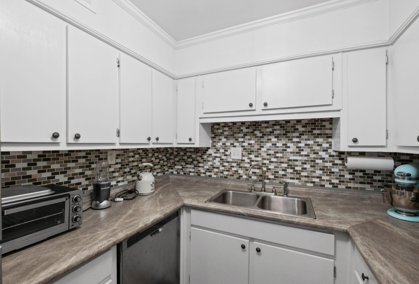 Kitchen featuring white cabinetry, stainless steel dishwasher, ornamental molding, tasteful backsplash, and light countertops