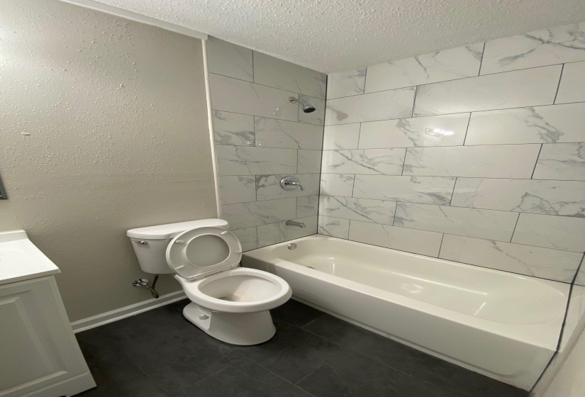 Bathroom featuring vanity,  shower combination, a textured ceiling, a textured wall, and dark tile patterned floors