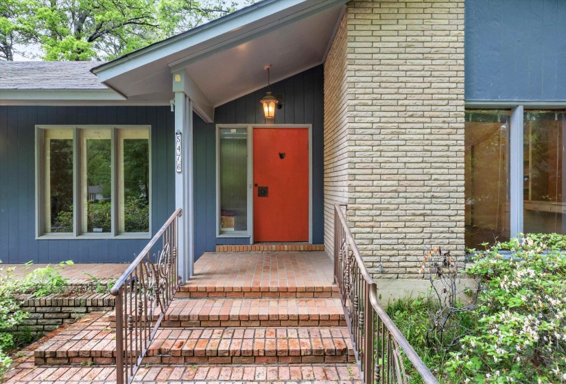 Welcoming covered Front Entrance showcasing classic mid-century modern design with clean lines, a bold statement front door, and brick steps with iron railing leading to a private, inviting entry framed by natural surroundings.