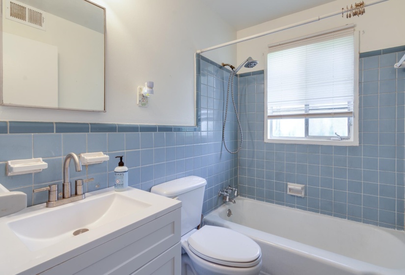 Bathroom featuring a white vanity with integrated sink, coordinating toilet, a bathtub with shower fixture, a window with horizontal blinds, and blue square wall tiling with a decorative border