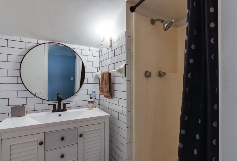 Bathroom featuring subway tile walls, a white vanity with louvered doors, a round black-framed mirror, and a shower with a beige surround
