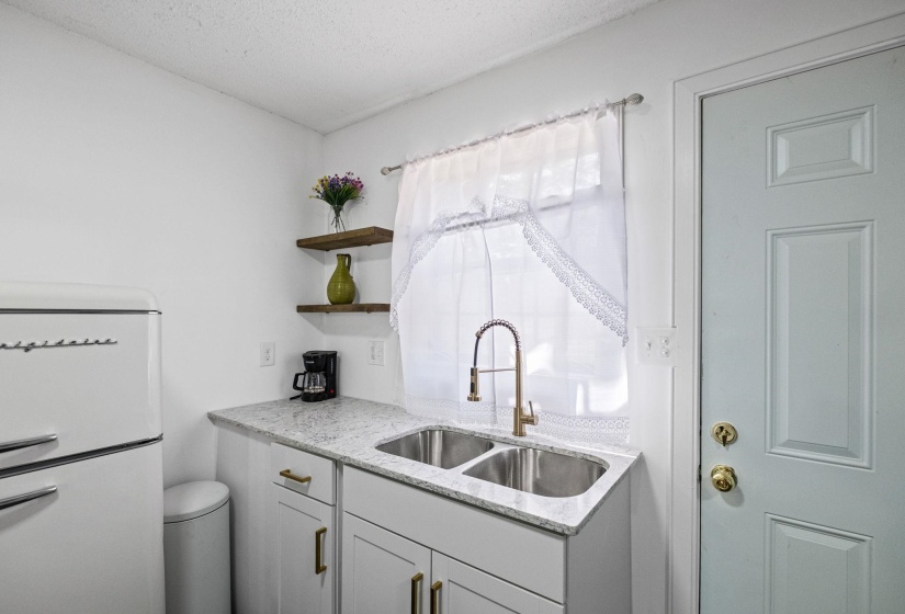 Kitchenette featuring a double basin stainless steel sink with a brass-finish pull-down faucet, light granite-style countertop, and white cabinetry with brass-finish hardware