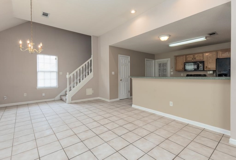 Vaulted ceiling great room featuring tile flooring, a chandelier, and a prominent staircase with white balusters