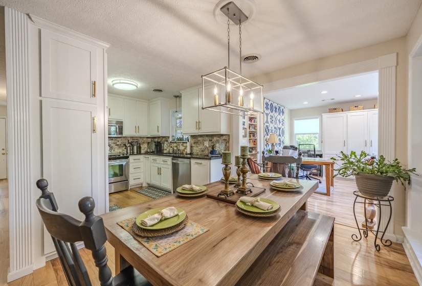 Open-concept dining area with wood-finish flooring, featuring a contemporary linear chandelier, white built-in cabinetry, and a clear sightline into the kitchen with stainless steel appliances