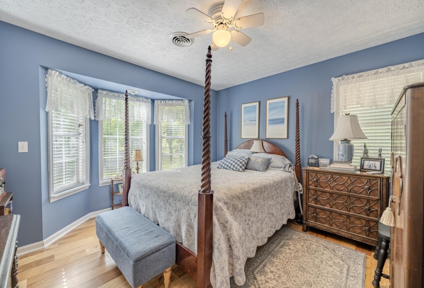 Bedroom featuring light wood-finish flooring, a bay window with three individual windows, a ceiling fan with integrated lighting, and light blue wall tones