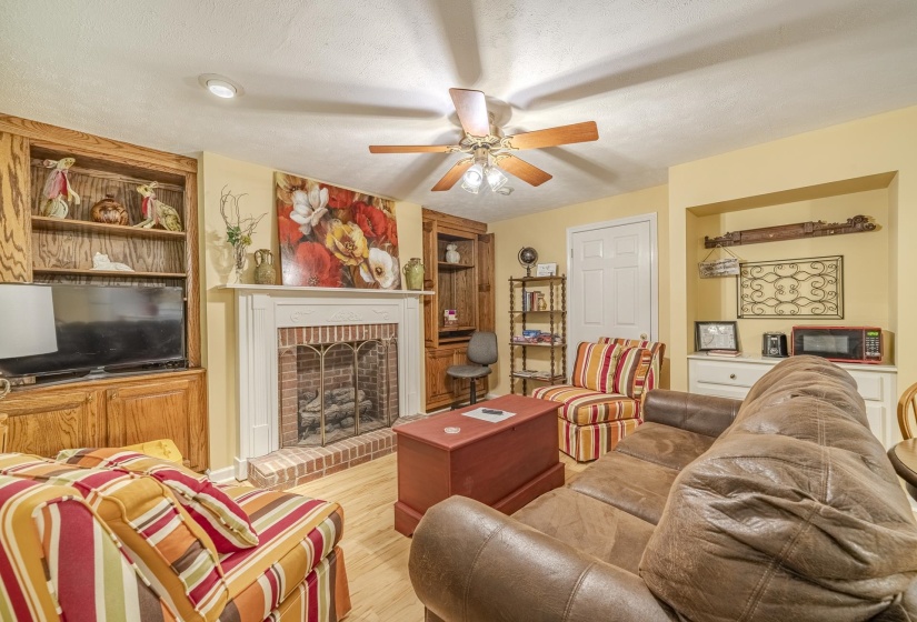 Living area featuring a brick fireplace with a white mantel, wood-finish flooring, recessed lighting, a ceiling fan, and built-in wood cabinetry