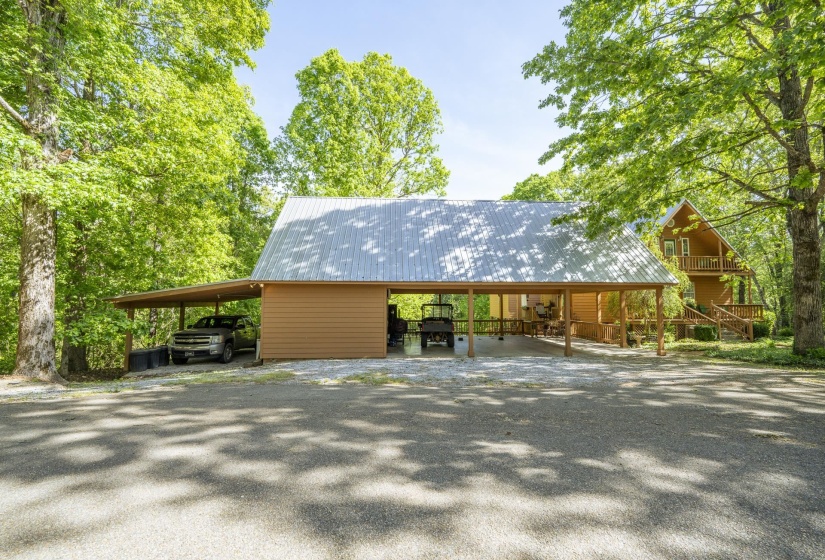 Expansive gravel drive with a carport structure featuring a corrugated metal roof and wood posts, complementing a distant A-frame style residence with a balcony and exterior staircase