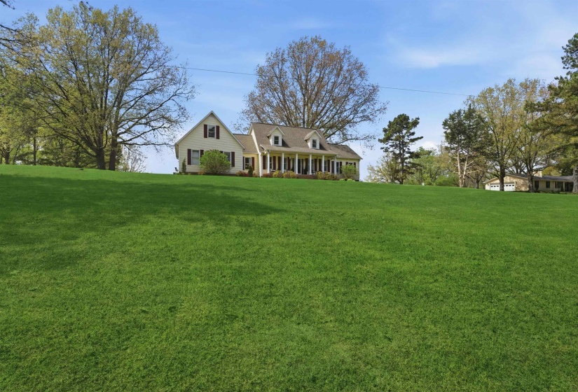 Expansive front lawn with mature trees framing a traditional-style home featuring a covered front porch with white columns, dormer windows, and dark shutters
