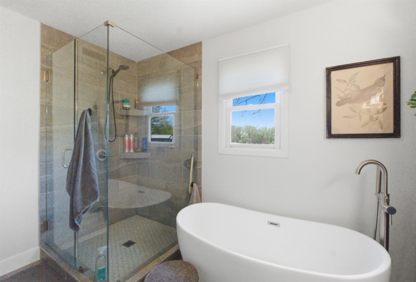 Freestanding soaking tub with floor-mounted faucet, glass-enclosed shower with tiled walls and hexagonal floor, and a window with roller shade