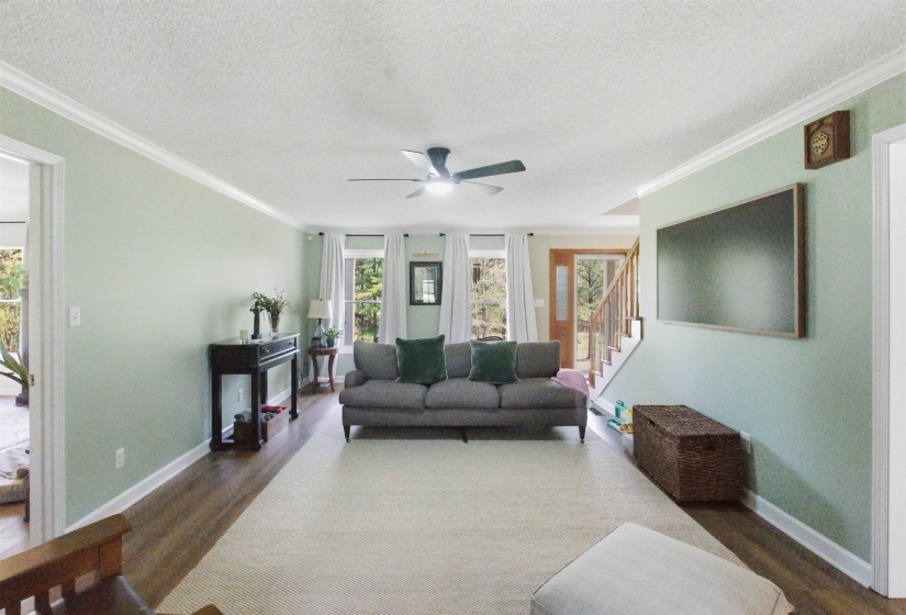 Open-concept living space featuring wood-finish flooring, a ceiling fan with integrated lighting, multiple windows with white window treatments, and white crown molding