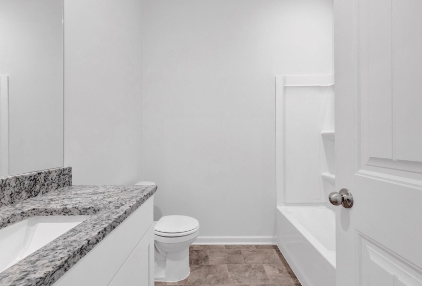Bathroom featuring a white vanity with an integrated sink, a speckled countertop, a toilet, a white bathtub with a built-in shower surround, and patterned flooring