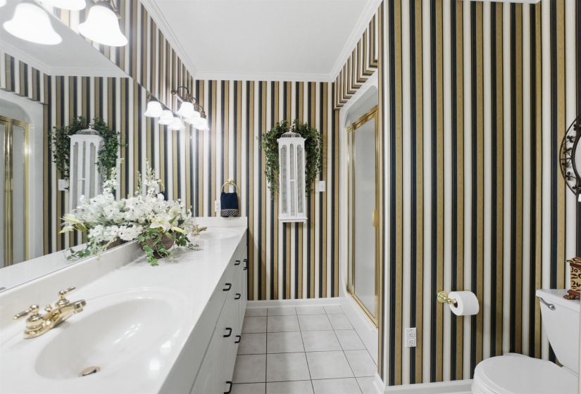 Bathroom featuring striped wallpaper, a white vanity with an integrated sink, polished brass fixtures, an overhead light bar, and a framed shower enclosure