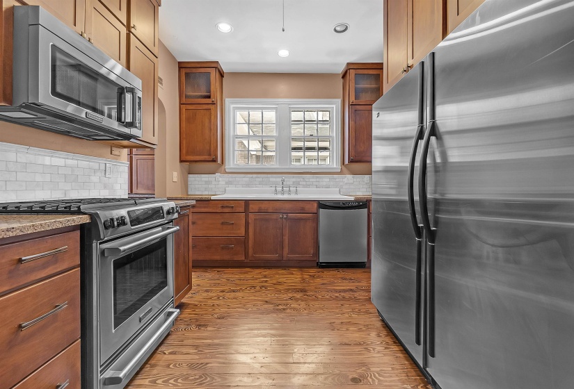 Kitchen featuring wood-finish flooring, wood cabinetry with glass-front accents, stainless steel appliances, a subway tile backsplash, and recessed lighting