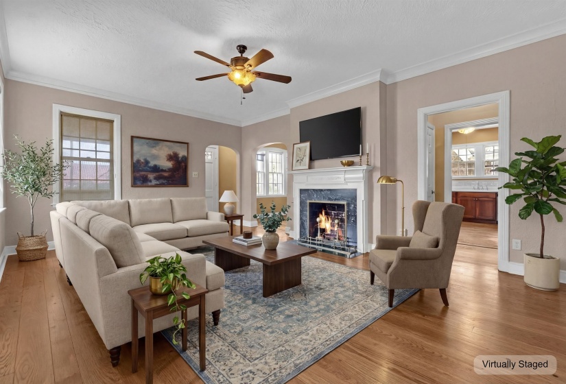 Living room featuring wood-finish flooring, a white mantel fireplace with a dark stone surround, crown molding, and a ceiling fan with integrated lighting