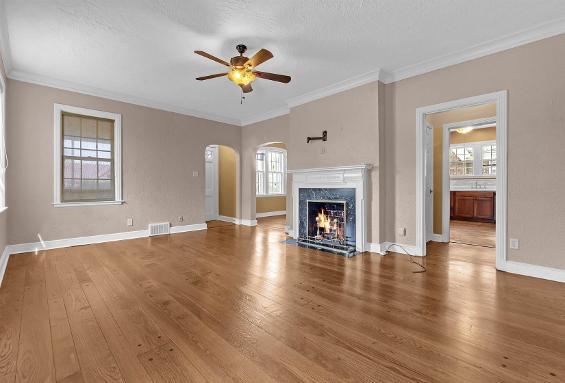 Living area featuring wood-finish flooring, a fireplace with a white mantel and dark stone surround, crown molding, a ceiling fan with integrated lighting, and multiple windows