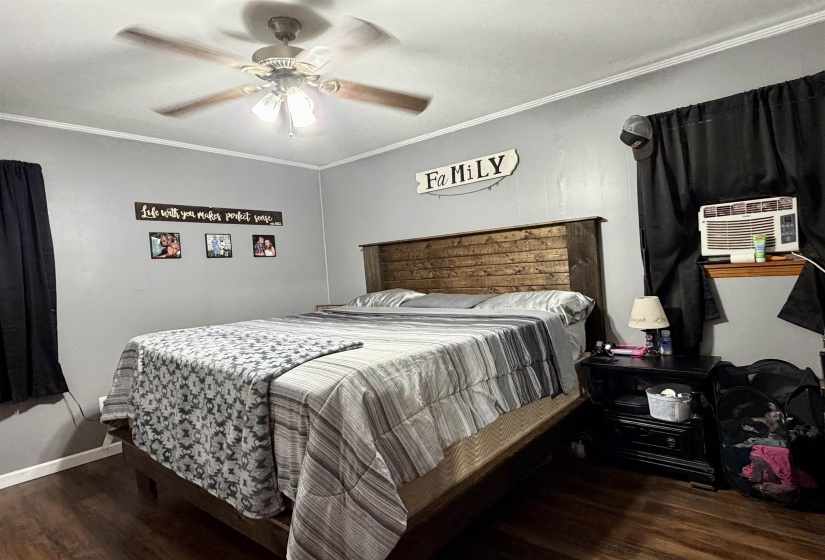 Bedroom featuring a ceiling fan with integrated lighting, dark wood-finish flooring, light gray wall paint, white crown molding, and a window with a built-in air conditioning unit