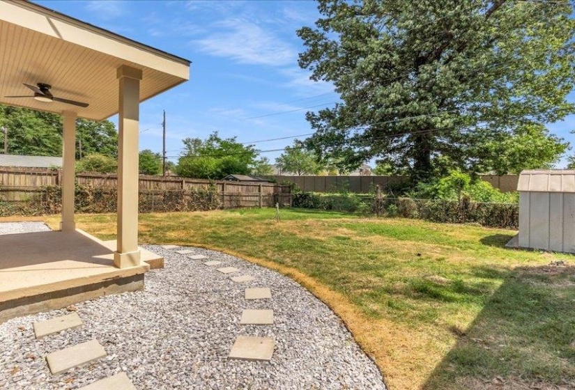 Expansive backyard featuring a covered patio, ceiling fan, gravel path with stepping stones, mature trees, and a storage shed