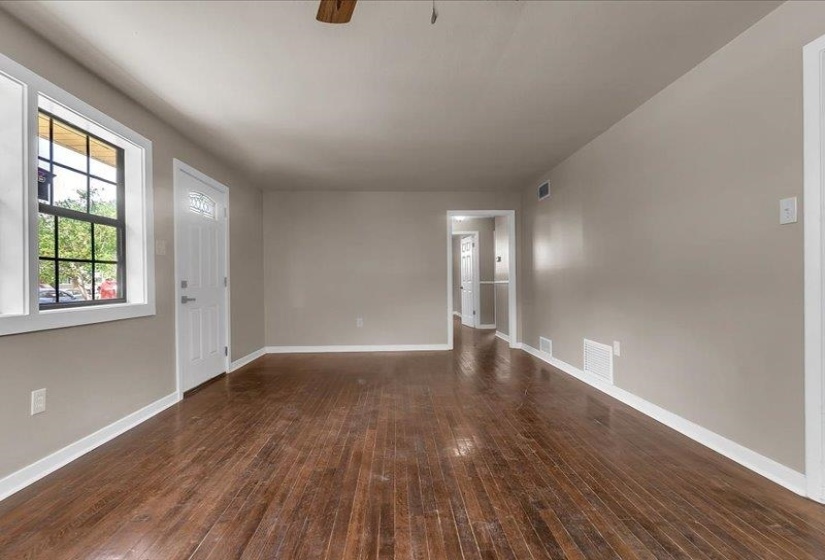 Spacious room with wood-finish flooring, neutral-toned walls, white trim, a white entry door with glass inserts, and multiple windows providing natural light