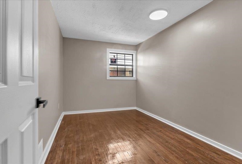 Inviting room with wood-finish flooring, neutral-toned walls, a window with dark framing, and a ceiling-mounted light fixture