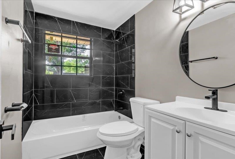 Modern bathroom featuring a black marbling-effect tile surround for the tub and shower, a white vanity with integrated sink, a circular framed mirror, and matte black fixtures