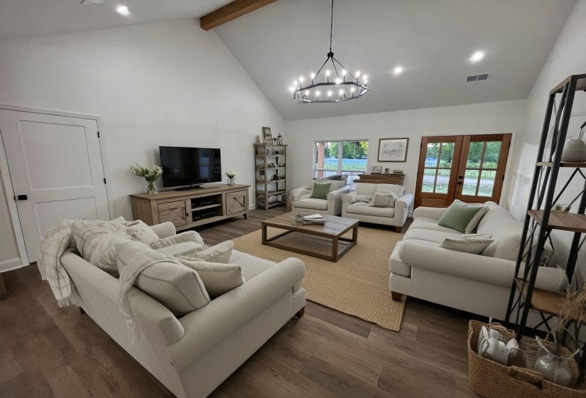 Spacious living area featuring a vaulted ceiling with an exposed wood beam, wood-finish flooring, a large circular chandelier, and light wood French doors