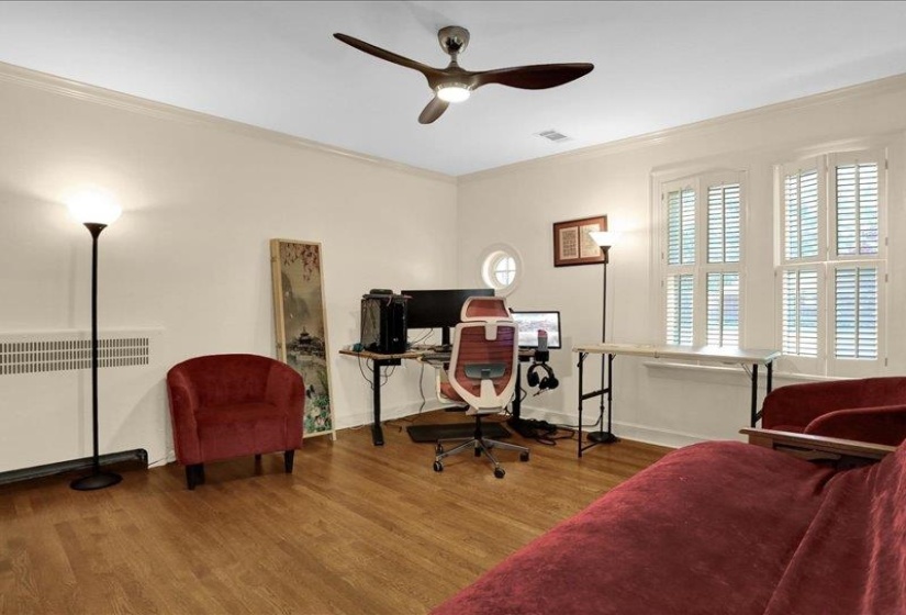 Hardwood flooring throughout featuring crown molding, two windows with plantation shutters, a round porthole window, and a ceiling fan with integrated lighting. NICE!