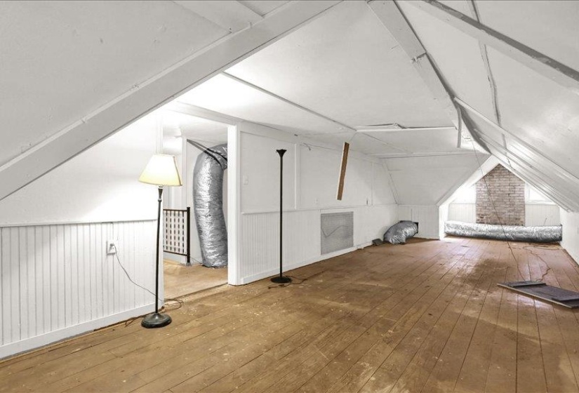 Spacious attic featuring wood-finish flooring, white paneled walls, and exposed ceiling beams.