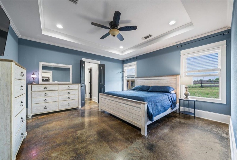 Spacious bedroom featuring stained concrete flooring, a tray ceiling with recessed lighting and a ceiling fan, and multiple windows offering natural light