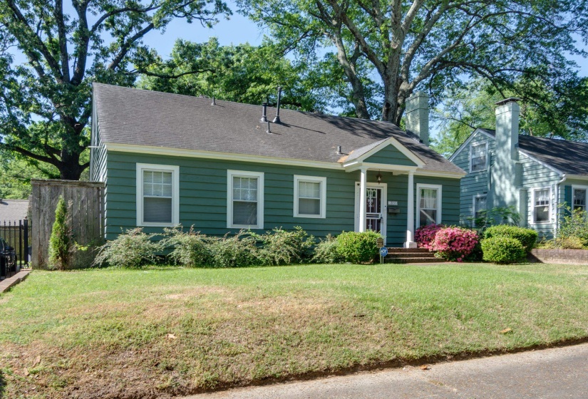 Traditional-style residence featuring green siding, a covered entry porch, white-trimmed windows, and mature perimeter landscaping