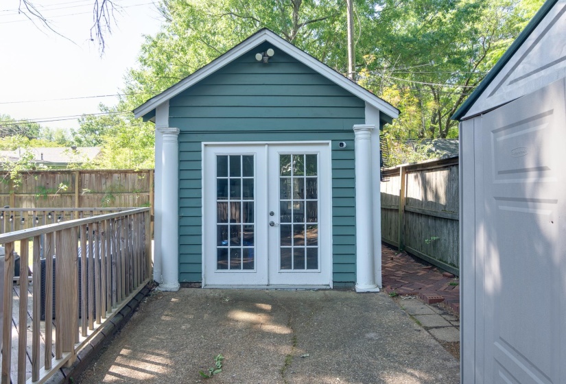 Blue exterior shed with gabled roof, double French doors, concrete patio, and fenced perimeter