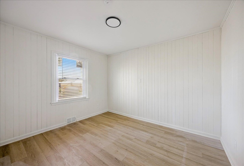 Light-filled room featuring a single window with blinds, white vertical paneling on two walls, smooth white walls, wood-finish flooring, and a flush-mount ceiling light fixture