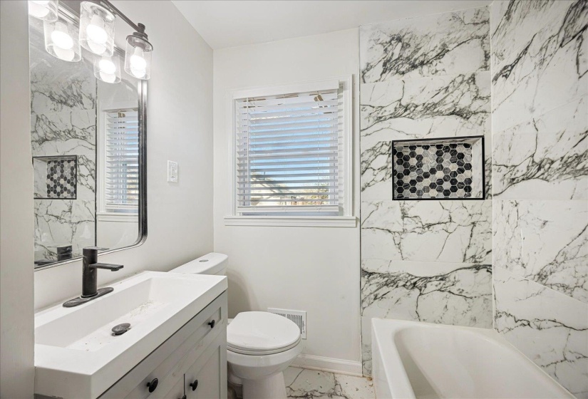 Bathroom featuring a white ceramic bathtub, large-format marbled tile surround with an integrated mosaic-tiled niche, a light wood-finish vanity with an integrated sink, and a black matte faucet