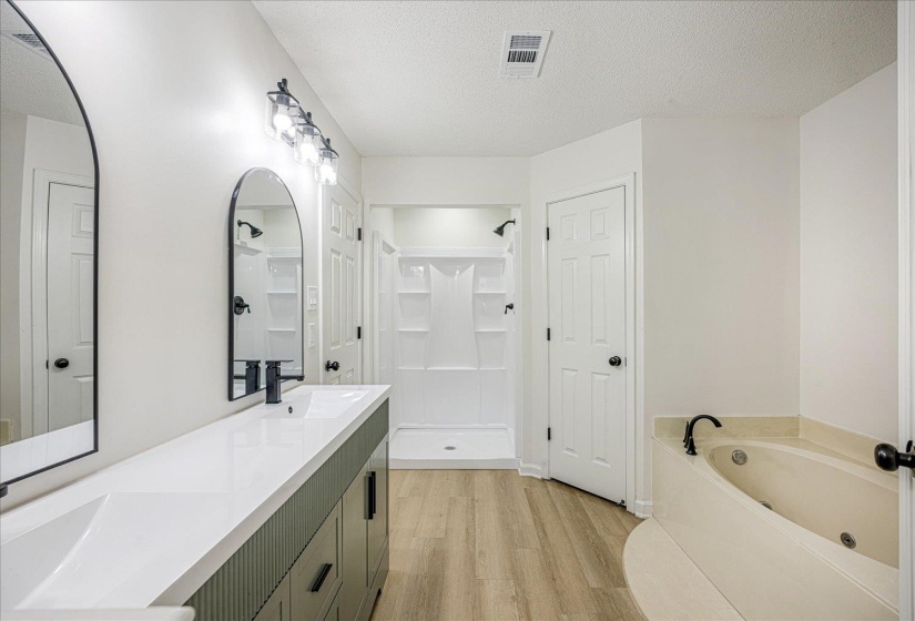 Bathroom featuring a dual vanity with a white countertop and matte black fixtures, arched mirrors, a walk-in shower with a white surround, a built-in jetted tub, and wood-finish flooring