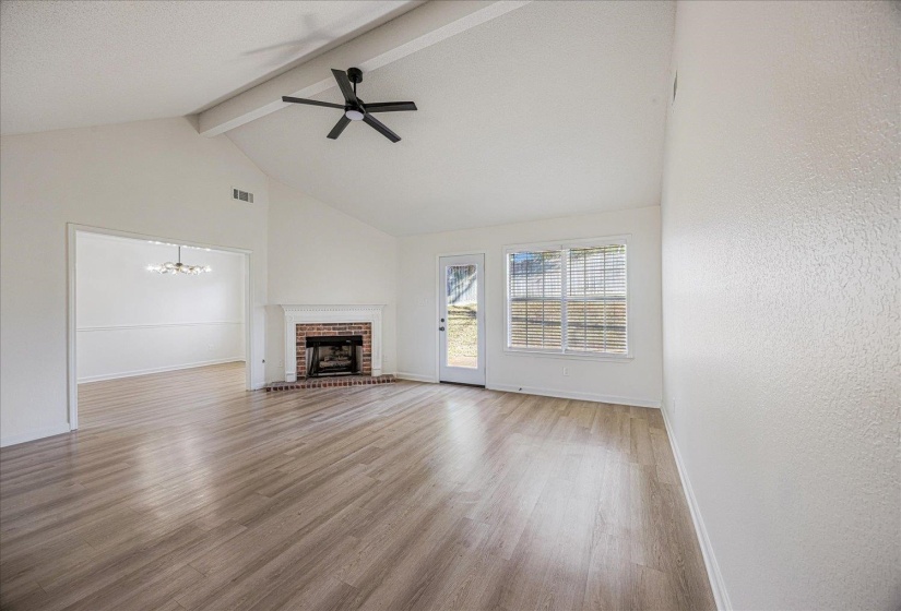 Spacious living area featuring a vaulted ceiling with exposed beams, wood-finish flooring, and a brick fireplace with a white mantel