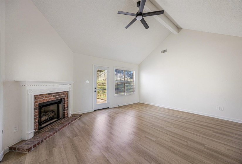 Spacious interior featuring a vaulted ceiling with exposed beam, wood-finish flooring, a brick fireplace with a white mantel, and a glass-paneled door