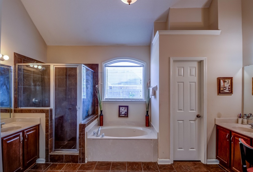 Spacious bathroom featuring an arched window above a built-in soaking tub, a separate glass-enclosed shower with tiled walls, and dual vanity areas with under-sink cabinetry