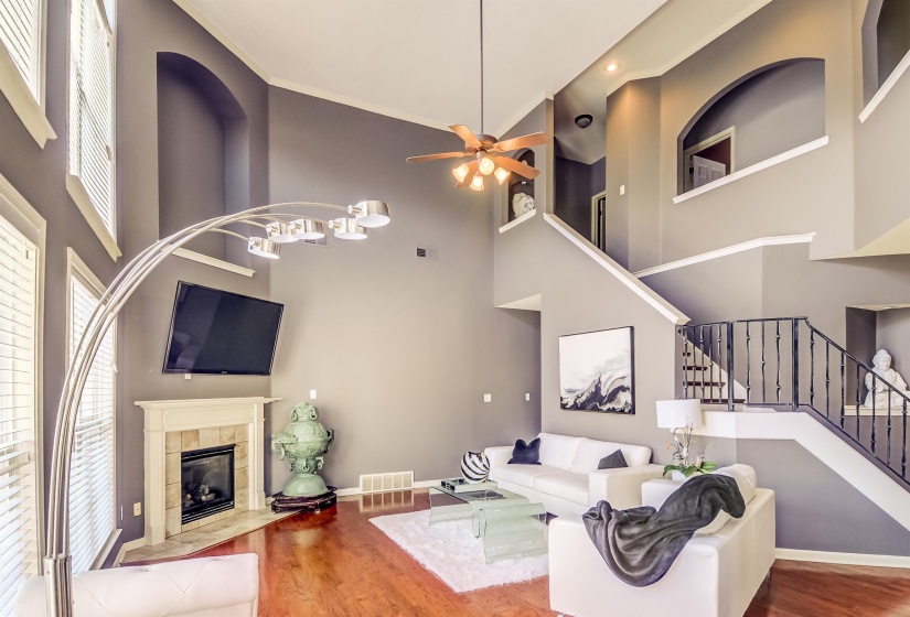 Two-story living area featuring wood-finish flooring, a corner fireplace with a white mantel, and a multi-level ceiling with a ceiling fan