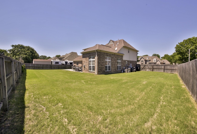 Expansive grassy backyard featuring a full perimeter privacy fence, brick exterior with large windows, and a paved patio area