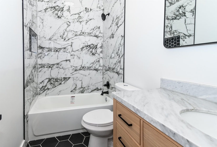 Bathroom featuring a bathtub with a tiled surround, a built-in shower niche, a toilet, a vanity with a light wood finish, and a stone-finish countertop with an integrated sink