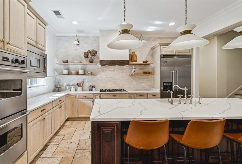 Spacious kitchen featuring an expansive island with a waterfall countertop, integrated stainless steel appliances, light-toned cabinetry, and natural stone tile flooring